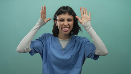 Woman nurse wearing scrubs raises hands and grimaces while making eye contact in teal studio;...