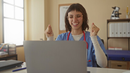 Woman wearing blue scrub top and stethoscope clapping hands over laptop at desk in a clinic building with anatomical chart behind; pride success.