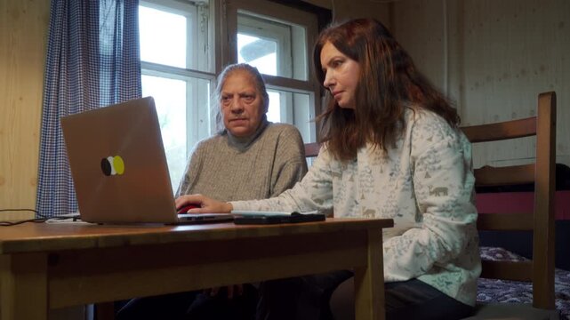 A middle-aged daughter shows her mother how to use laptop and red mouse on a wooden table near a window with checkered curtains. Both women appear focused and serious while engaging with the computer