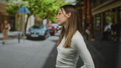 Young woman standing with hand on hip on a busy city street, face turned to camera in a white long...