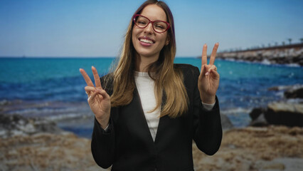 Woman smiling and showing two peace signs with both hands at a sunny beach pier wearing red glasses and black blazer; joy vacation fun.