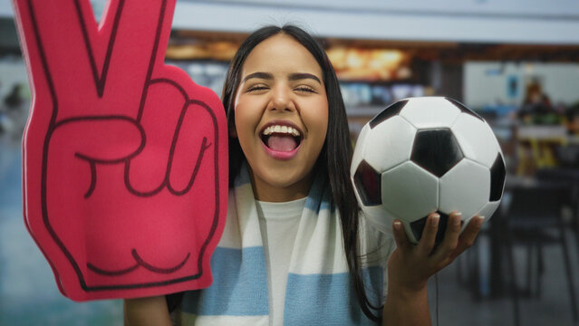 Woman smiling indoors holding soccer ball and foam glove in restaurant setting showcasing youthful energy and vibrant fan spirit.
