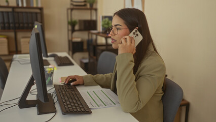 Latin woman in office working on computer call holding reports wearing glasses and jacket.