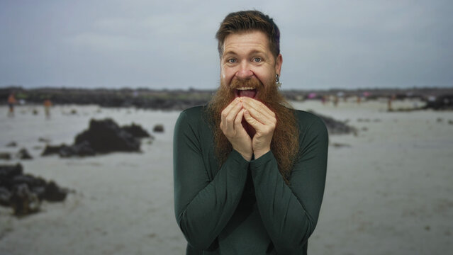 Young caucasian man in green shirt biting nails on sandy beach with rocky shore and wet sand under overcast sky; anxiety.