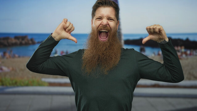 Man with beard giving thumbs down with open mouth under bright sunlight at beach overlooking sea; rejection.