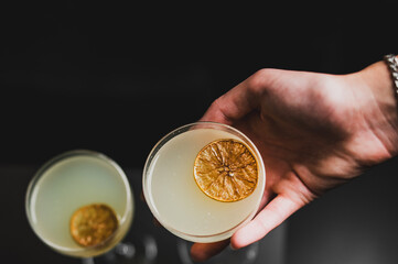 Fototapeta premium A hand holds a yellow cocktail in a coupe glass, garnished with a dried citrus wheel, against a dark background with a second blurred drink nearby