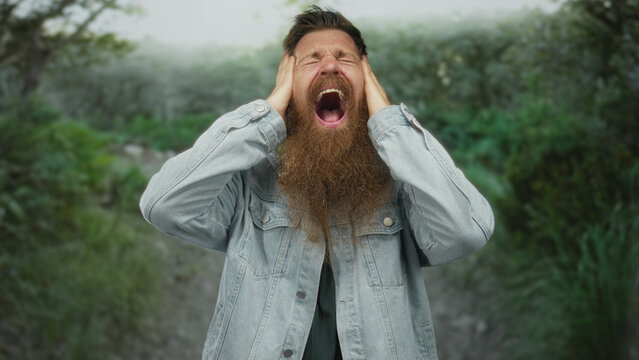 Young man with long beard and denim jacket screams while holding his head among green park trees; surprise.