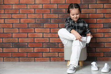 Little girl trying on different shoes near brick wall indoors. Space for text