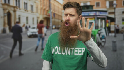 Young caucasian man in green volunteer shirt smiles and points finger on city street; community...
