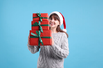 Obraz premium Smiling woman in Santa hat with many gift boxes on light blue background. Christmas and New Year celebration