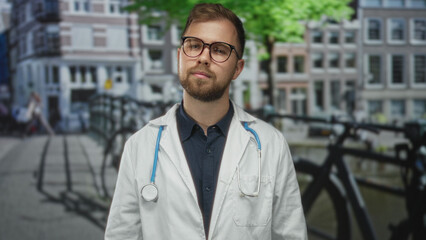 Man doctor wearing white coat and stethoscope standing on street by canal and building facades in amsterdam; urban healthcare thoughtful.