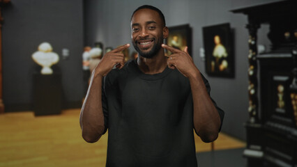 Young african american man points finger to smile in art gallery indoors with framed paintings and sculptures in view; pride confidence.