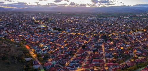 Twilight aerial panorama of Korçë with glowing streetlights, dense neighbourhoods and mountain silhouettes under a fading sunset sky.