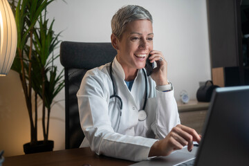 Mature female receptionist talking on phone in clinic while sitting and looking on pc monitor
