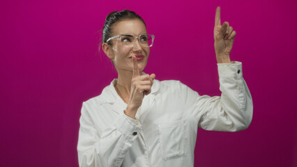 Young hispanic woman scientist in white lab coat points finger upward in pink studio; inspiration curiosity innovation discovery.