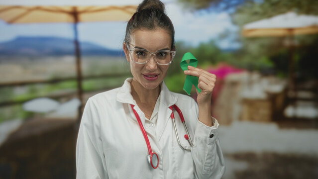 Woman doctor smiling holds green ribbon with stethoscope on restaurant terrace; supportive hope health awareness.