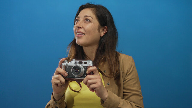 Middle aged hispanic woman holding vintage camera with bare hands in blue professional studio environment; curiosity.