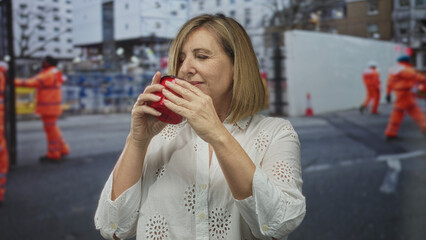 Fototapeta premium Woman holding red cup near nose while standing on street at construction site wearing white blouse and focusing on drink aroma; serenity.