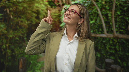 Woman with red glasses points finger upward in forest, smiling and wearing white shirt and olive blazer; optimism.