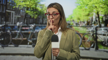 Fototapeta premium Blonde young woman holding microphone, finger to lips for silence on street in amsterdam wearing blazer and red glasses during on camera report; secrecy.