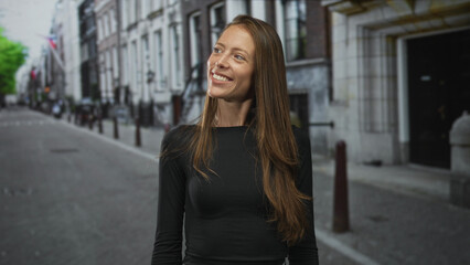 Fototapeta premium Woman smiling, teeth visible, looking up while wearing black long sleeve shirt on a city street lined with stone buildings; urban happiness.