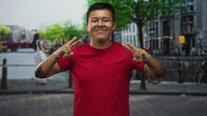 Hispanic man showing peace sign with right hand by amsterdam canal in a red shirt and smiling broadly; happy travel leisure.