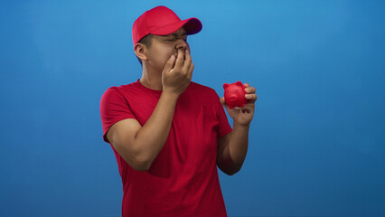 Young hispanic man biting fingernails and holding a small red piggybank in a blue studio; financial...