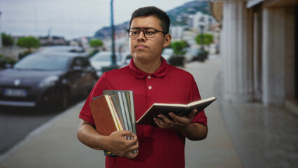 Young hispanic man wearing glasses holds an open book and a stack of textbooks while reading with a puzzled expression on a city street near parked cars; confusion.