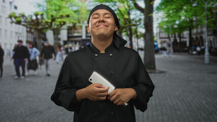 Fototapeta premium Young man chef in black uniform holding a cleaver to his chest, eyes closed and smiling while standing on a cobbled street; pride in craft.