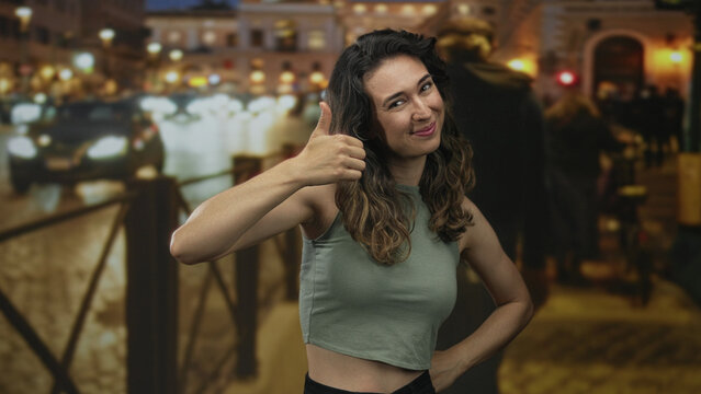 Woman showing thumbs-up on city street at night under warm streetlights with blurred traffic lights and passing cars; optimism.
