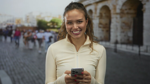 Woman smiling holding smartphone in front of roman coliseum in italy outdoors surrounded by tourists capturing the historic ambiance on a sunny day.