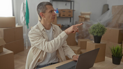 Man in casual attire working on laptop surrounded by cardboard boxes and plants in new apartment living room setting with natural light streaming in through windows.
