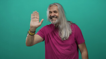 Man waving hand in studio with teal backdrop, long silver hair and visible rings smiling broadly; friendly welcome joy.