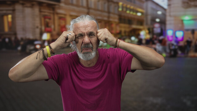 Man covering ears with fists in street, wearing magenta shirt and gray pulled back hair, grimacing against loud traffic and crowd noise; noise annoyance.
