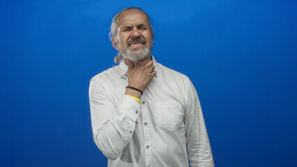 Man with long gray hair and beard holding throat in blue studio wearing white shirt; discomfort health concern.
