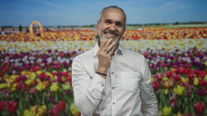 Man with gray long hair and beard smiling with hand on chin in a colorful tulip field wearing a white shirt; contentment.