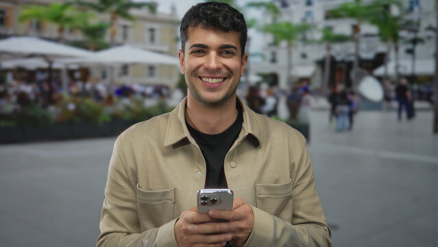 Man inspects smartphone while holding plaza backdrop, young hispanic man previews photo on phone as male model poses in outdoor setting feeling happy.