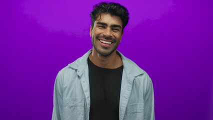 Fototapeta premium Man standing smiling with visible face and tilted head in studio portrait against vibrant purple wall; joy.