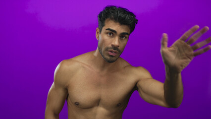 Fototapeta premium Young hispanic man with muscular bare chest points fingers downward in purple studio; invitation.