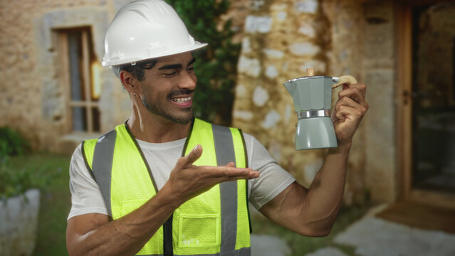 Young hispanic engineer wearing hardhat and neon vest points finger to coffee pot at building entrance; pride.