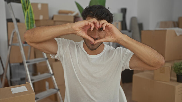 Man forms heart shape with hands amid moving cardboard boxes in new home building interior; expressing love. - Powered by Adobe