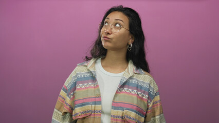 Fototapeta premium Woman points finger up and looks up in a pink studio, wearing round glasses and patterned jacket over a white tee; curiosity.