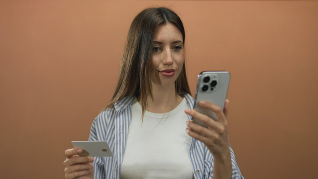 Woman holding creditcard and smartphone against orange studio wall with focused gaze; uncertainty decision.