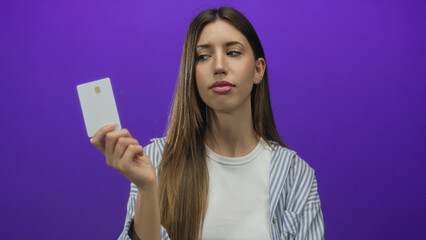 Fototapeta premium Young woman holds creditcard against purple studio wall with a serious expression and determined stare; confidence.
