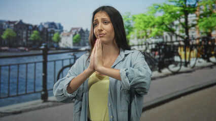 Fototapeta premium Young hispanic woman with hands clasped in prayer and closed eyes on amsterdam street by canal railing, wearing denim shirt; hope plea.