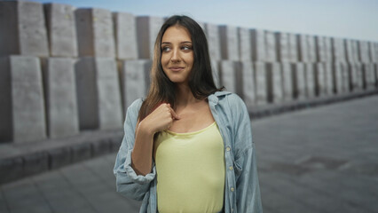 Fototapeta premium Hispanic young woman in yellow tank top and denim shirt points finger to chest on a city street by stacked concrete blocks, smiling slightly; confidence.