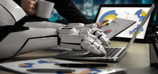 A robotic arm works on a laptop amidst a business setting, representing the fusion of technology and productivity in a modern office.Noogenesis