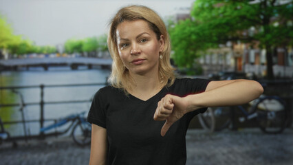 Blonde woman shows thumbs down gesture on street by canal and bicycle railing in city scene; disapproval dissent.