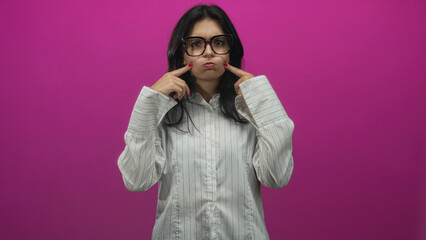 Young hispanic woman pushes cheeks outward with both index fingers in bright pink studio setting; toothache discomfort.