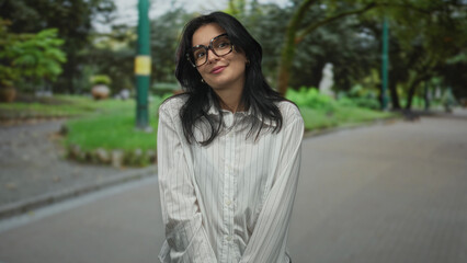Young hispanic woman wearing glasses tilts head with a slight smile on urban street; quiet contemplation.